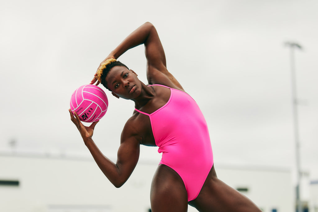 Ashleigh Johnson, a Black Olympic water polo player poses with a hot pink water polo ball and wears a hot pink one piece swimsuit