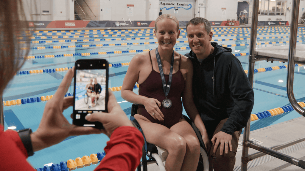 Mallory Weggeman sits wearing a gold medal and her hand on her pregnant belly next to her husband Jay in front of a pool. Both are smiling.