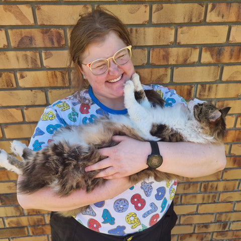 Photo of MB, holding Frank the cat who is cradled like a baby and touching her face with his paws. MB is wearing black shorts, glasses, and a white teeshirt with colourful numbers on it.