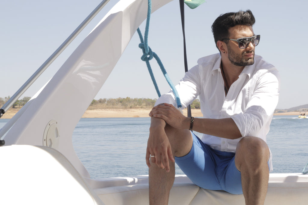 a man wearing white shirt, with blue shorts and sunglesses with the sea on the background 