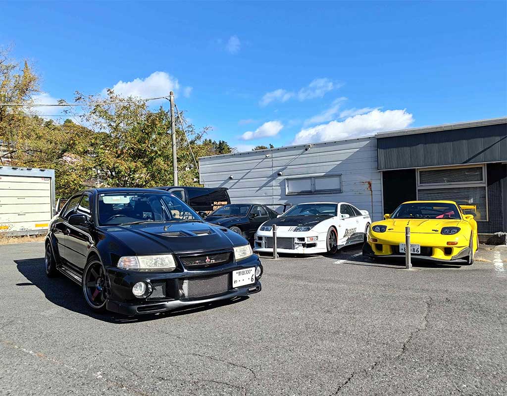 An RX7 FC, Evo, and RX7 FD in front of a cafe under Mt.Akina in Japan