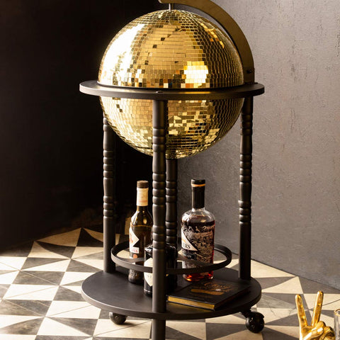 Lifestyle image of the Black & Gold Disco Ball Drinks Trolley Cart displayed on a geometric floor in front of a black wall, styled with bottles on the bottom shelf of the trolley.
