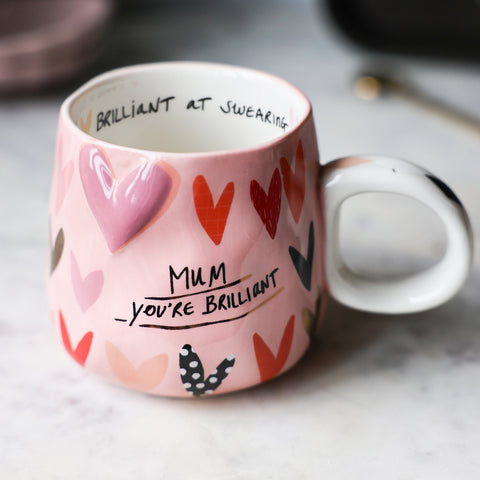 Lifestyle image of the Mum, You're Brilliant Mug displayed on a table with kitchen accessories in the background.