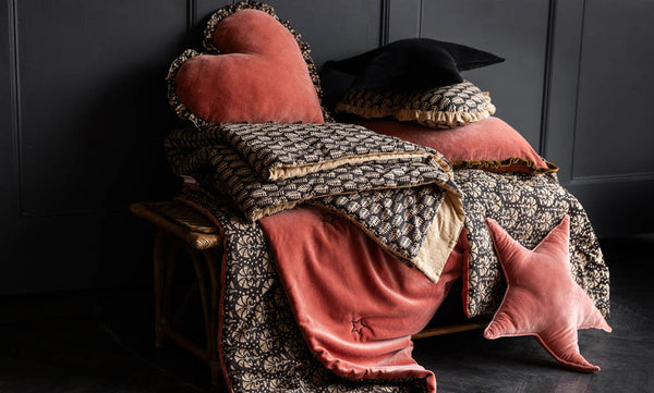 A bench layered with pink and black cushions and throws in velvet and block printed materials, styled against a dark panelled wall.