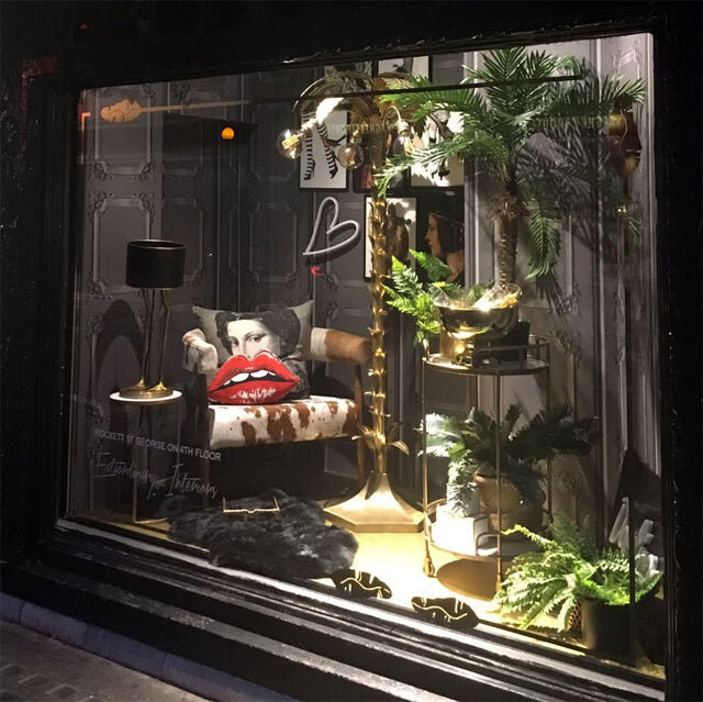 A shop window displaying an eclectic mix of objects, from a cow-hide armchair to a lips cushion, black lamp and golden palm tree