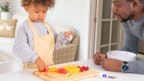toddler boy chops colourful fruit with a doddl toddler knife, watched by his dad