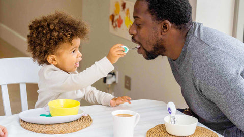 A toddler boy with curly hair feeds his dad with a doddl toddler spoon at the meal table