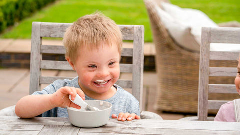 Toddler boy sitting at a garden table eating yoghurt with a doddl toddler spoon, with big smiles