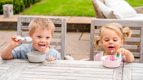 Toddler boy and toddler girl happily sitting at a garden table eating pink yoghurt