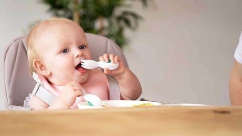 Baby sits at wooden table eating with doddl baby cutlery