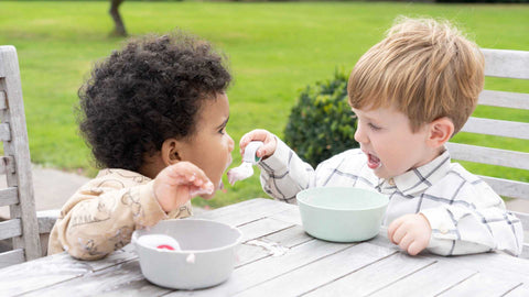 Two toddler boys sitting at a garden table with spoons and bowls, one is feeding the other for fun