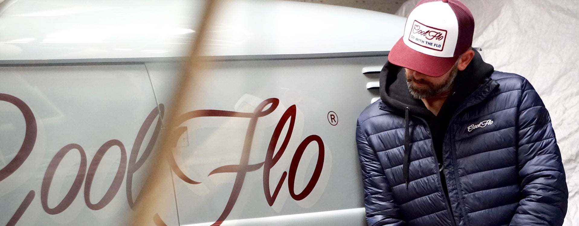 Male model wearing a Cool Flo navy puffer jacket and burgundy and white cap, standing by a grey Cool Flo VW bus.