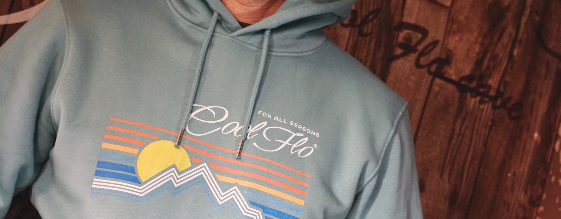 Close-up of model wearing a light teal hoody with a graphic linear mountain and sun 'For All Seasons' full colour design printed on the front.