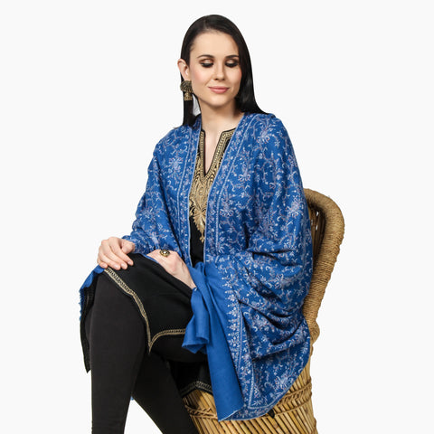A model sitting on a. chair wearing a blue color Pashmina shawl with white embroidery around it