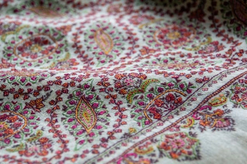 A closeup picture of a Pashmina Shawl with a white base fabric and intricate paisley embroidery in multiple colors including red and green.