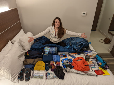 Girl sits on a bed with an array of backpacking gear