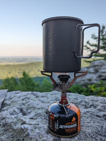 A small metal pot with handles sits on top of a backpacking stove.  There are mountains in the distance.