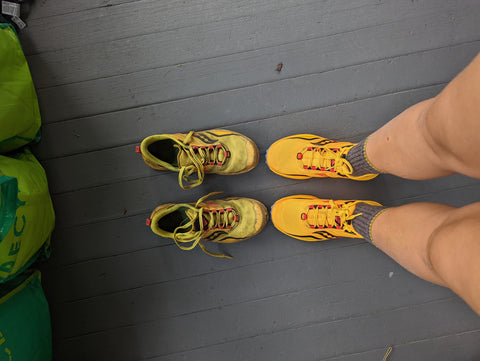 Looking down at a hiker's legs and shoes.  She is wearing a new, bright yellow pair of trail runners.  Next to her feet are and old beat up pair of the same shoes.