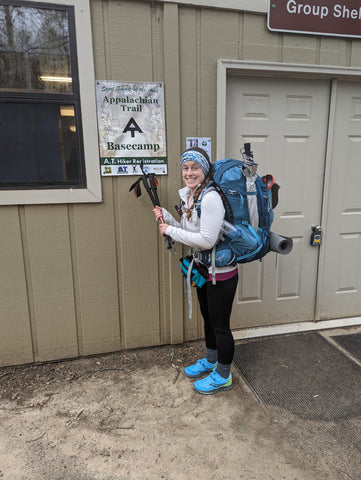 A young woman with a large backpack and trekking poles stands in front of a sign for AT thru-hiker basecamp