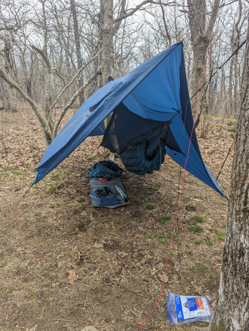 A blue hammock hangs under a blue tarp.  There is a backpack and some supplies on the ground underneath.