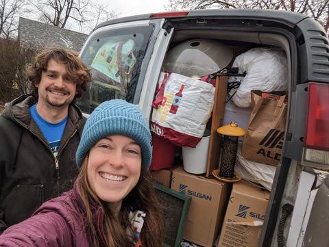 A young man and woman smile in front of van that is completely full of boxes and bags.