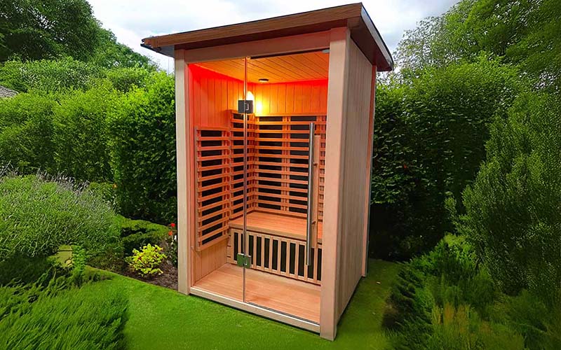 A stylish Insignia Outdoor Infrared Sauna set against lush greenery, showcasing the sleek design with tempered glass door, warm internal lighting, and premium Canadian Hemlock construction.