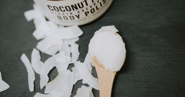 A spoon of exfoliating coconut body polish sits on a table alongside shredded coconut