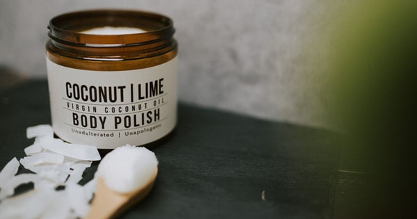 A jar of coconut and lime body polish sitting opened on a table. A spoon of the body polish is on the table with shredded coconut scattered around.