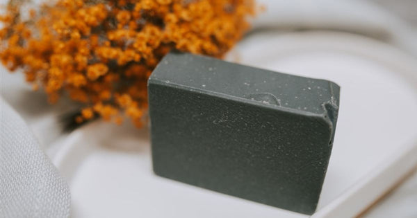 A black handmade soap bar sitting on a tray in front of a bunch of dried flowers