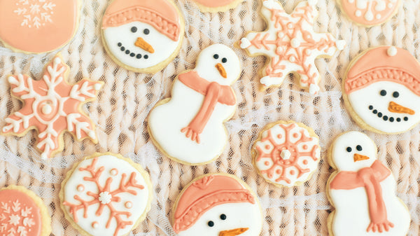 This image is a variety of Holiday frosted sugar cookies in the shapes of snowmen or snow people and snowflakes. They are decorated in white and coral icing and they are laying in a cute arrangement on a soft peach colored piece of looped fabric, that looks like a woven blanket.