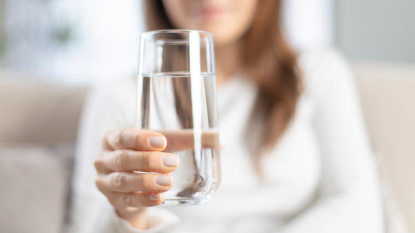 This is an image of a clear glass of water being held out to the front of the image by a woman's hand.  You can see the woman blurred out in the background, she is wearing a white long-sleeved shirt and you cannot see her full face.