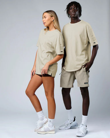 One female model and one male model both wear the LSKD oversize tee in taupe. Male model wears taupe LSKD shorts also and female model wears black booty shorts. Both standing with white trainers