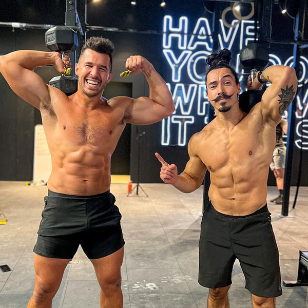 Two men standing with one dumbbell on shoulder in gym topless