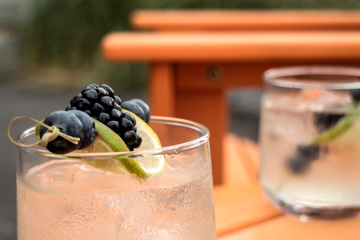 Two refreshing cocktails with berries and lime garnishes on a POLYWOOD outdoor table, set against a blurred background of an orange outdoor chair, suggesting delightful outdoor entertaining.