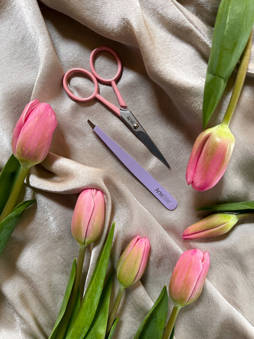 A pair of tweezers and a pair of scissors on a beige velvet background. There are pink tulips surrounding the frame.