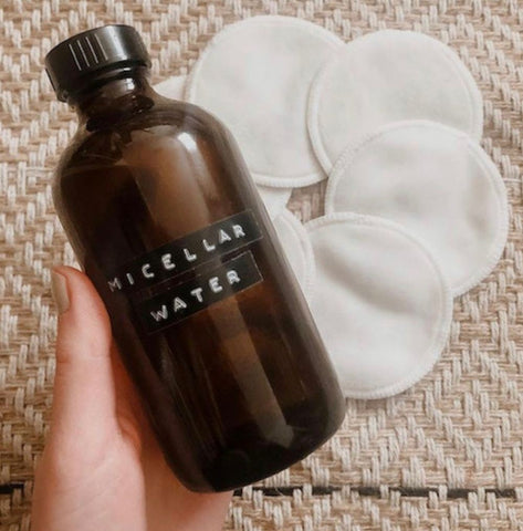A hand holding a brown bottle labelled "micellar water". It features a beige background with white cotton pads.