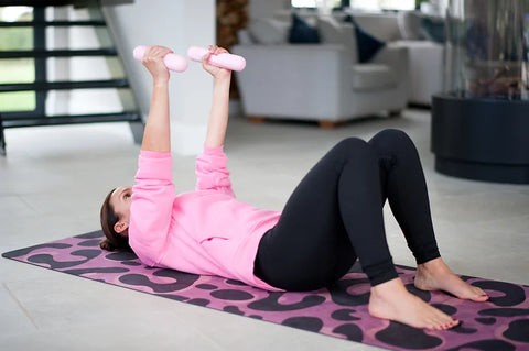 Lady lies on her back on leopard print yoga mat holding hand weights above her shoulders in chest press