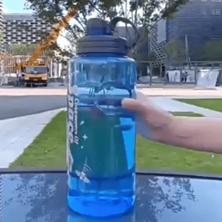 Quelqu'un fait tourner rapidement une grande Gourde sport bleue | Space™ de Mon Univers Sport, partiellement remplie d'eau. En plein air, entre bâtiments, herbe et arbres, idéale pour s'hydrater au quotidien.