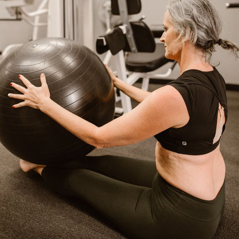 Senhora praticando ioga ou pilates, sentada no chão com a perna esticada e uma bola em cima