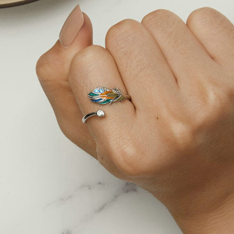 Um anel de prata 925 com uma pena colorida e pedras brilhantes na parte superior, visto no dedo em zoom de mão feminina fechada.