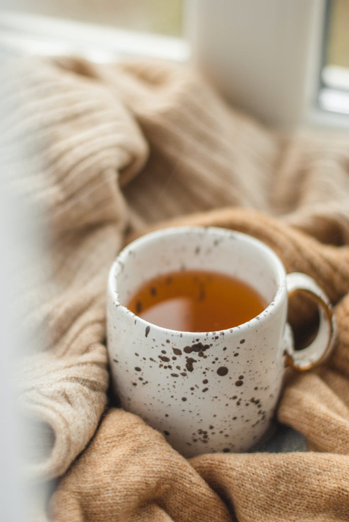 White ceramic mug with black speckles filled with amber liquid surrounded by light brown blankets