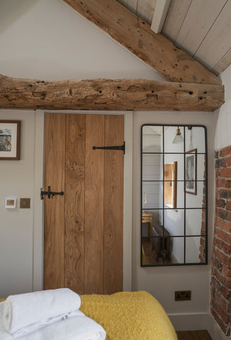 Ledge & brace door with From The Anvil's Beeswax T hinges & thumblatch, on a white wall with exposed oak beams above, and a patterned mirror to the side. A blurred bed with folded towels in the foreground and a reflection of a black radiator, wooden nightstand, and wooden window shutters in the mirror.