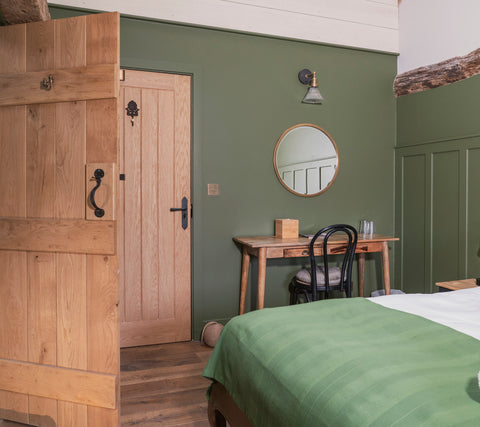 Open ledge & brace door with black hardware in a bedroom with a wood floor, green walls, and a green double bed, a wooden desk with a black chair and a round mirror on the wall above it.