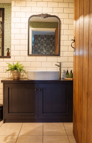 A ledge & brace door with Pewter hardware opening into a bathroom with white wall tiles, a blue cabinet, a large white ceramic sink, a potted plant, and assorted glass bottles.