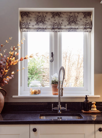 From The Anvil's Black Monkeytail window espag. on a white framed kitchen window with a patterned blind, black countertop, and white cabinets with black handles.