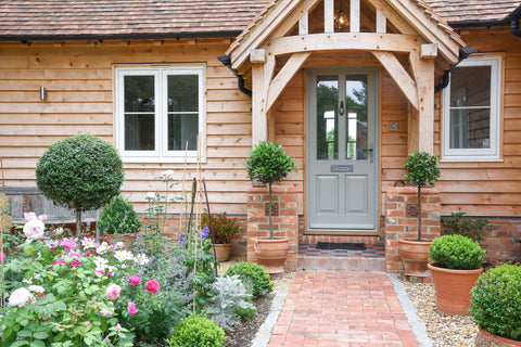 Sage green door with a Black From The Anvil door knocker, with a timber frame in front, covering a red brick path through a front garden filled with green plants.