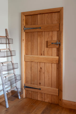 Wooden ledge & brace door with From The Anvil Pewter T hinges and thumblatch against a white wall in a room with a wooden floor and a rustic wooden ladder with wire mesh baskets attached.