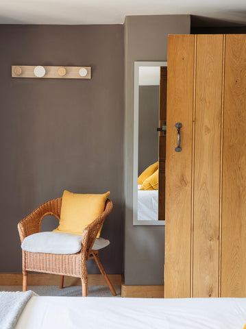 Wooden ledge & brace door with Pewter From The Anvil thumblatch, against a dark grey wall with a long mirror, and a wicker chair with an orange cushion.