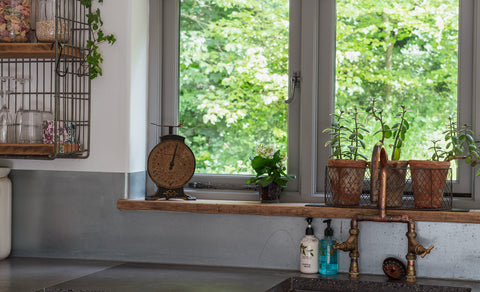 From The Anvil Pewter Peardrop window espag. handles on a grey framed window with a wooden sill filled with potted plants and an antique scale, with a shelving unit to the left, and a granite countertop with rustic copper taps below.