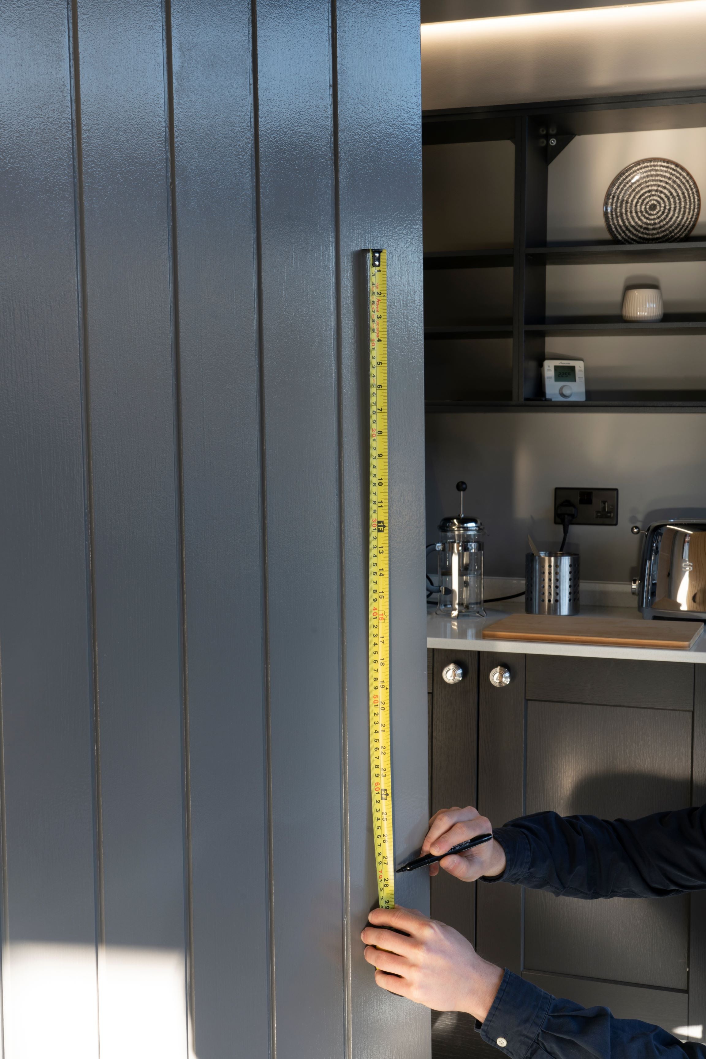 Person using a tape measure to measure the distance between two fixing points on a T bar pull handle on a grey panelled door.